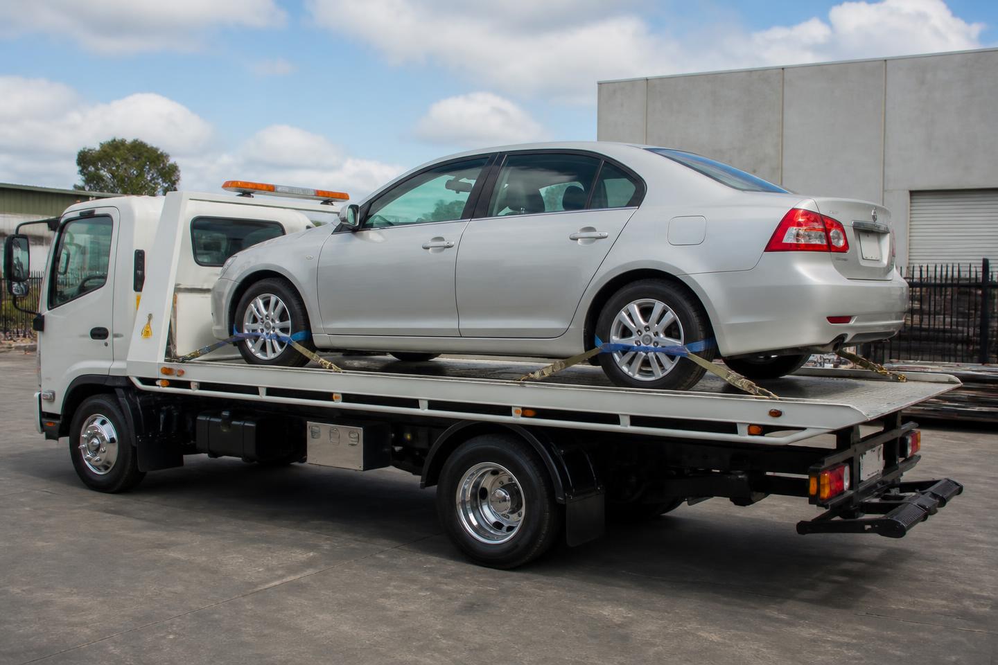 Silver sedan secured with over-the-wheel straps on the level tray of a vehicle transport truck in an Australian yard